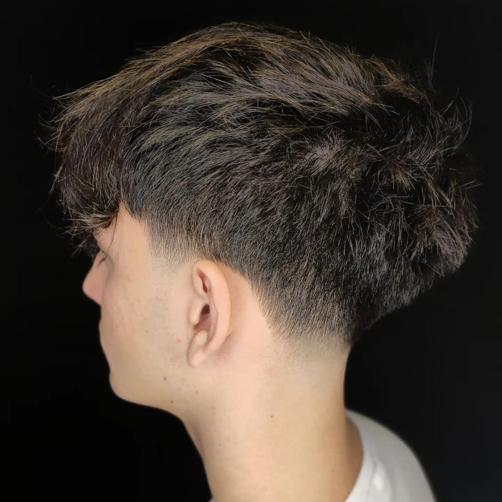 A side profile of a young man with thick, textured hair on top that gradually tapers down into a low, subtle fade near the nape of his neck. The top hair is left longer and slightly messy, adding volume and a casual look, while the sides feature a clean, soft blend into the fade. The dark background emphasizes the contrast between the longer top and the neatly tapered sides and back, creating a stylish and modern appearance. The overall look is effortless yet refined.