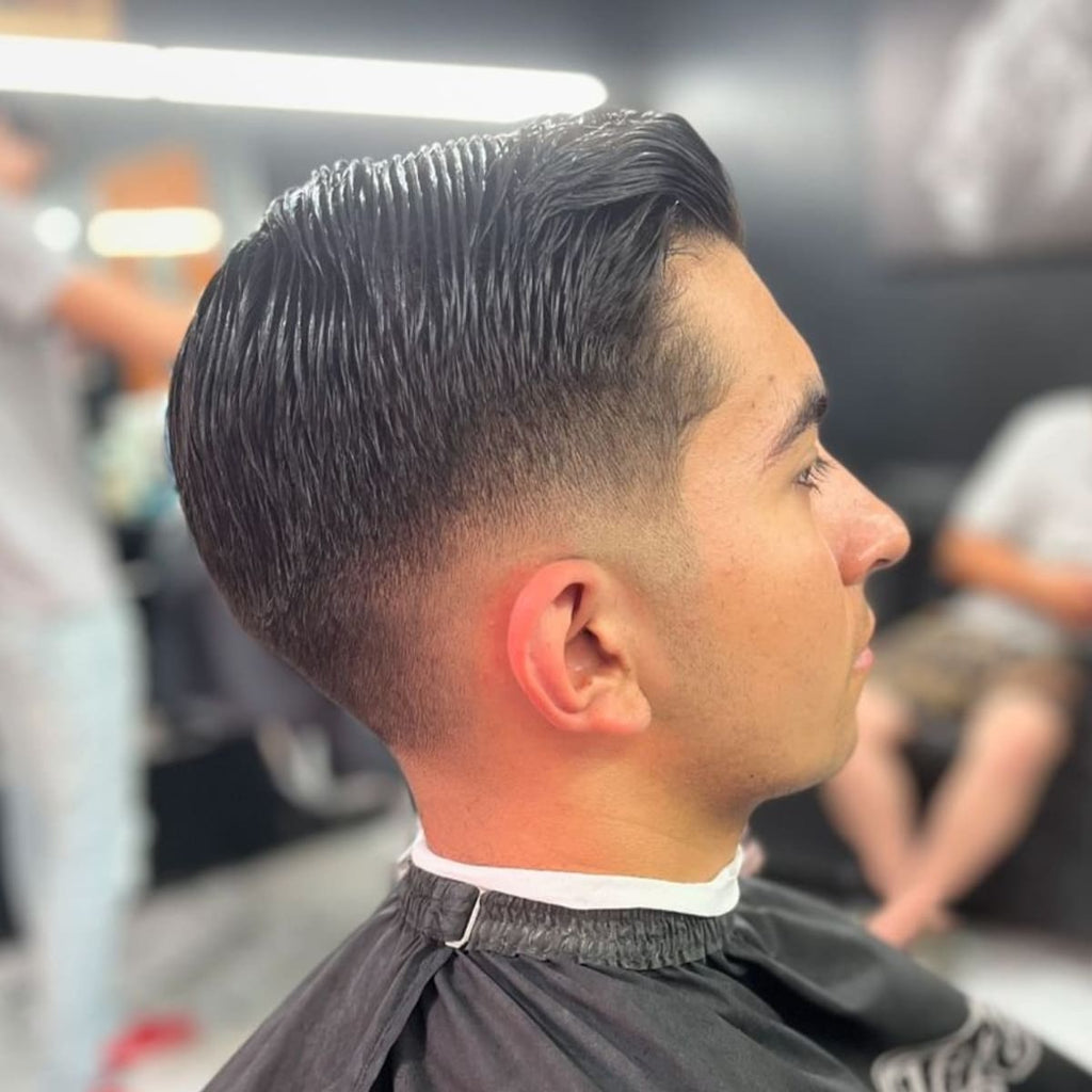 A side profile of a man sporting a comb over low fade haircut. The hair is neatly combed over to one side with a smooth and sleek finish on top, while the low fade seamlessly blends the hair down towards the neckline. The sharp, clean lines and tapered fade create a polished and well-groomed look, ideal for both casual and formal settings.