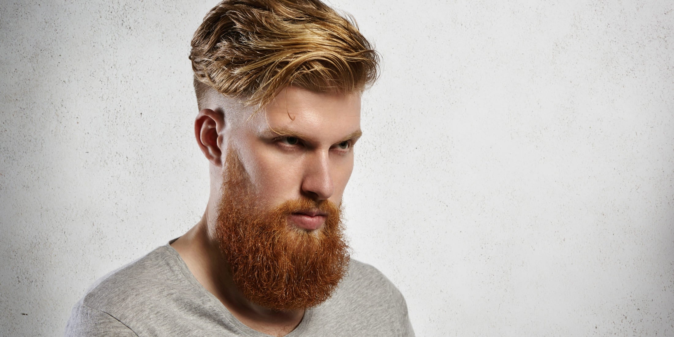 Portrait of a man with a blonde slick back undercut hairstyle and a full beard. The slicked-back top contrasts with the faded sides, showcasing a modern variation of the slick back undercut.