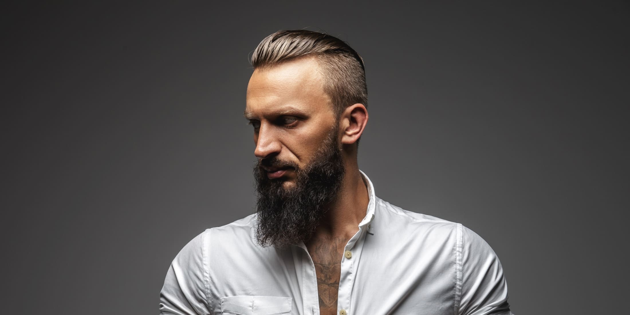 Man with a slick back undercut hairstyle and a long beard, featuring neatly slicked-back hair on top with closely cropped sides. This modern slick back undercut style is complemented by a stylish beard for a bold look.