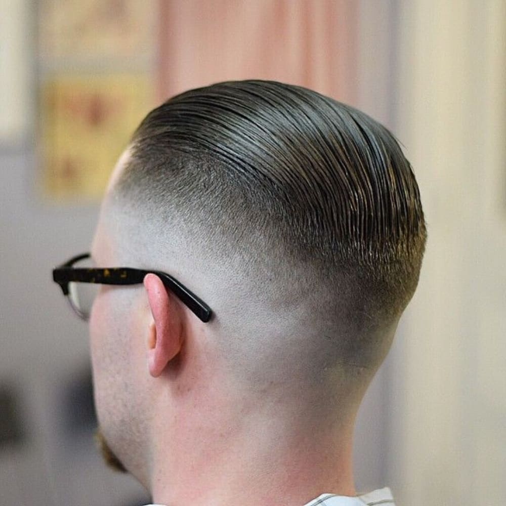 A man with a sleek, slicked-back hairstyle paired with a high fade. The hair on top is smoothly combed back, creating a polished and classic look. The high fade starts just above the temples and blends down to the skin, offering a stark contrast between the longer top and the closely cropped sides and back. The man is wearing black-rimmed glasses, and the view is from behind, highlighting the precise and clean lines of the high fade. The background is slightly out of focus, emphasizing the sharpness and detail of the haircut.