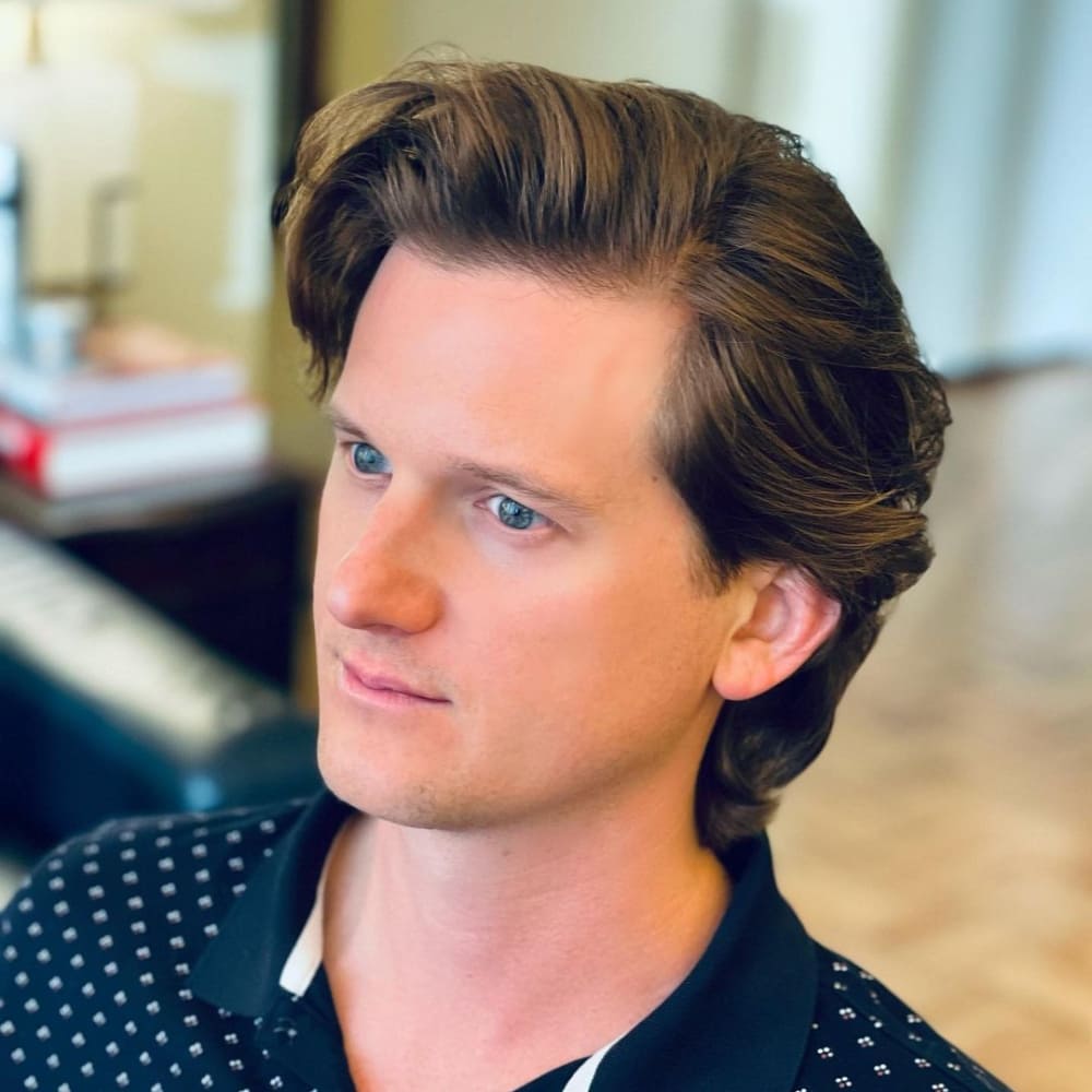 Man with straight hair styled in a bro flow haircut, featuring medium-length hair that flows naturally with a slight wave. He is wearing a black polo shirt with white accents and sitting indoors, looking off to the side, showcasing a relaxed and effortlessly cool look.