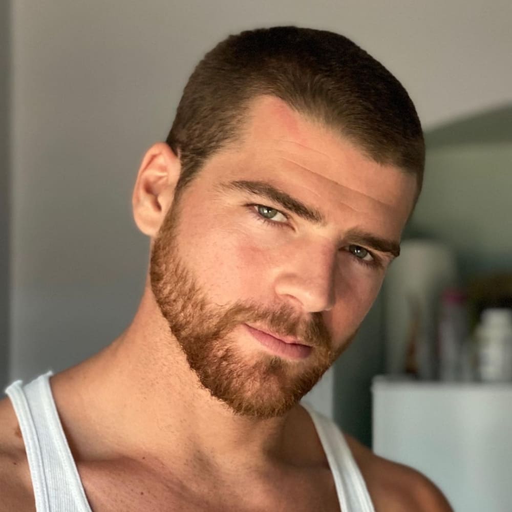 Man with straight hair sporting a clean, short buzz cut and a neatly trimmed beard, wearing a white tank top, and looking confidently into the camera with a background of a softly lit room.