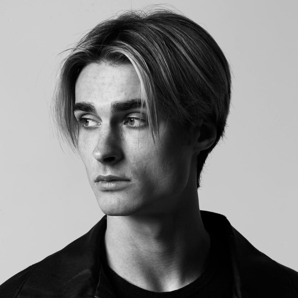 Young man with straight hair styled in a curtains haircut, featuring a center part with longer strands framing his face. He is wearing a black jacket over a dark shirt and looking off to the side, showcasing a modern yet retro-inspired look in a black-and-white photograph.