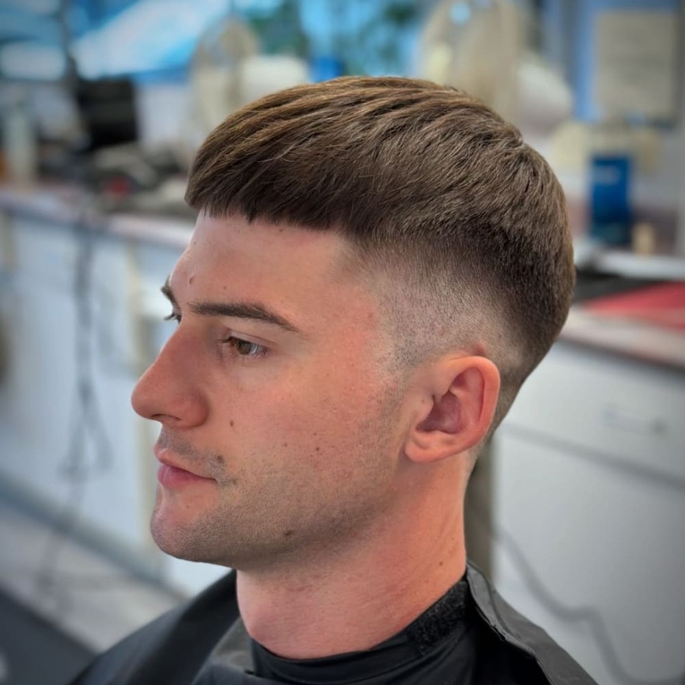 Man with straight hair sporting a French crop haircut, featuring short sides and back with a textured fringe. He is sitting in a barbershop chair with a black cape, looking to the side with a calm expression, showcasing the clean, modern style in a well-lit barbershop setting.