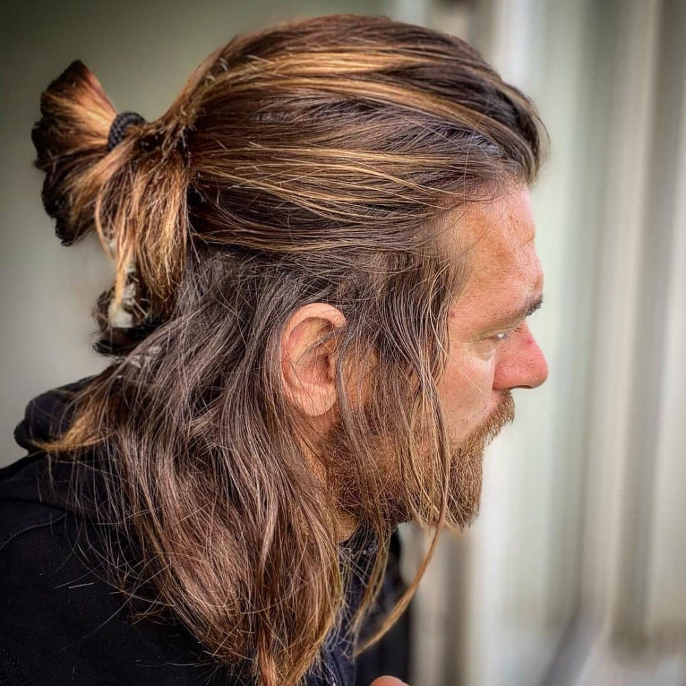 Man with straight hair styled in a half-up, half-down haircut, featuring the top section of his long hair tied back into a bun while the rest flows freely. He has a beard and is wearing a dark jacket, looking out a window, showcasing a relaxed and natural look with subtle highlights in a softly lit setting.