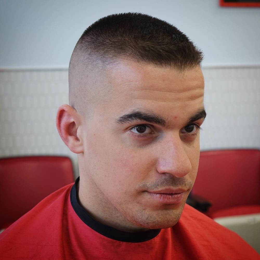 Man with straight hair featuring a high and tight haircut, sitting in a barbershop chair with a red cape. He has closely shaved sides and back with slightly longer hair on top, looking ahead with a calm expression.