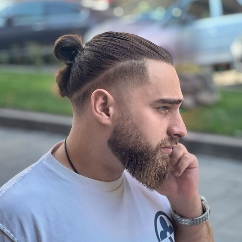 Man with straight hair styled in a man bun, featuring a sleek bun at the back with neatly shaved sides. He has a full beard and is wearing a light-colored T-shirt, standing outdoors and holding a phone to his ear, showcasing a modern and practical hairstyle in a casual setting.