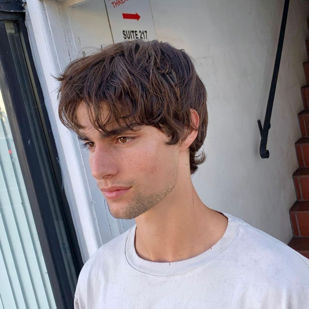 Young man with straight hair styled in a medium shaggy haircut, featuring layered, tousled hair that falls naturally around his face. He is wearing a light-colored shirt and standing outside near a storefront, showcasing a casual and effortless look with soft, natural waves.