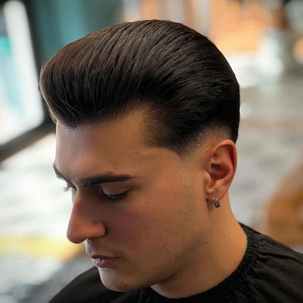 Man with straight hair styled in a pompadour haircut, featuring voluminous, slicked-back hair on top with short, faded sides. He is wearing a black cape and a small earring, looking down in a well-lit barbershop, showcasing a classic and polished look.