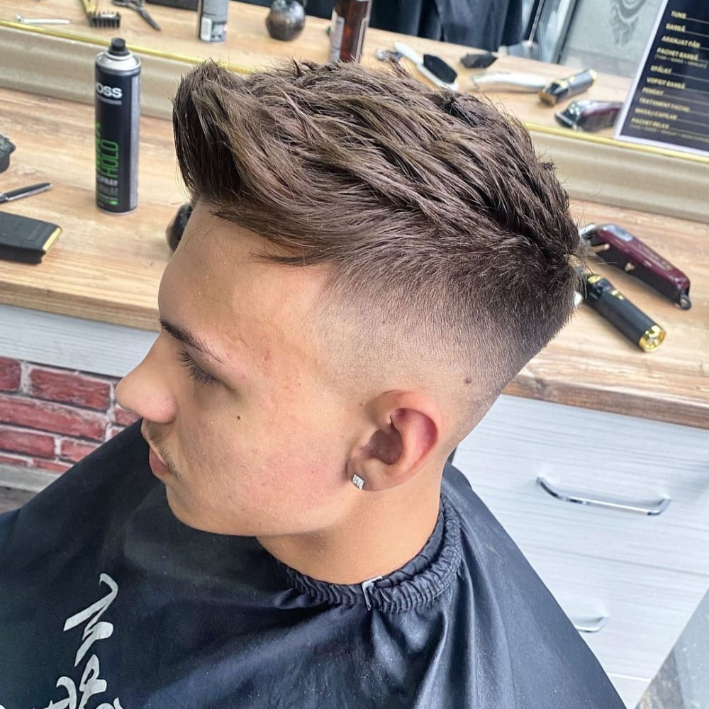 Young man with straight hair styled in a quiff haircut, featuring voluminous, textured hair on top with a high fade on the sides. He is wearing a black barber cape and an earring, sitting in a barbershop with grooming tools visible on the counter, showcasing a trendy and stylish look.