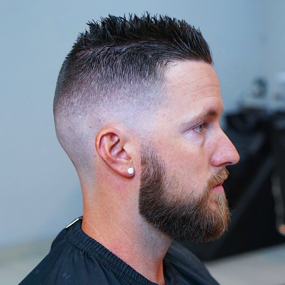 Man with straight hair styled in a short messy spikes haircut, featuring textured, spiked hair on top with a high fade on the sides and back. He has a well-groomed beard and is wearing a black barber cape, sitting in a barbershop, showcasing a modern and edgy look with an earring.