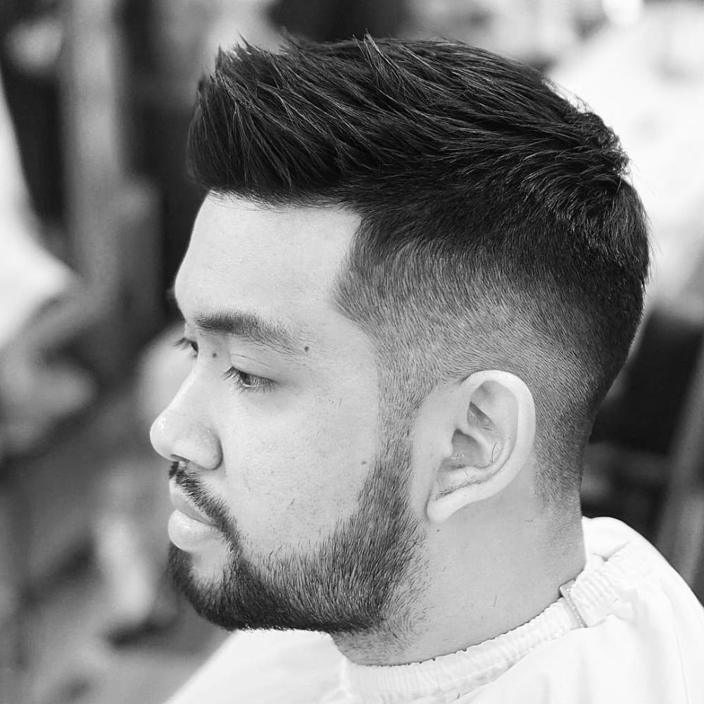 Man with straight hair styled in a short quiff haircut, featuring textured, upward-styled hair on top with short, faded sides. He has a neatly trimmed beard and is wearing a barber cape, sitting in a barbershop, showcasing a clean and modern look in a black-and-white photo.