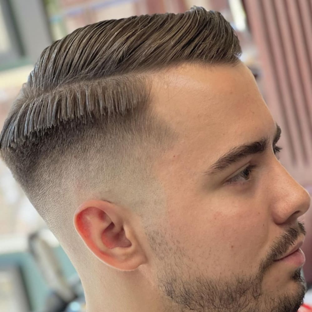 Man with straight hair styled in a side part haircut, featuring neatly combed, longer hair on top and short, faded sides. He has a well-groomed beard and is sitting in a barbershop chair, highlighting the clean and classic style in a modern setting.