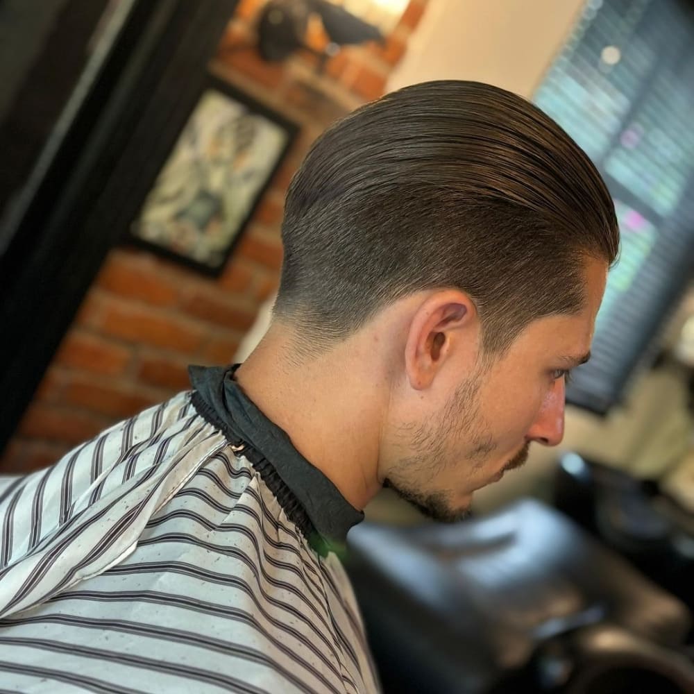 Man with straight hair styled in a slicked back haircut, featuring neatly combed hair that is sleek and glossy. He is sitting in a barbershop chair with a striped barber cape, showcasing a clean and professional look with a well-groomed beard, in a setting with exposed brick walls and soft lighting.