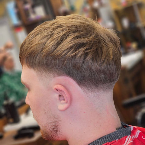 Man with straight hair styled in a tapered haircut, featuring longer hair on top with a gradual taper down the sides and back. He has a light beard and is wearing a red barber cape, sitting in a barbershop, highlighting the smooth transition of length in a relaxed and classic style.