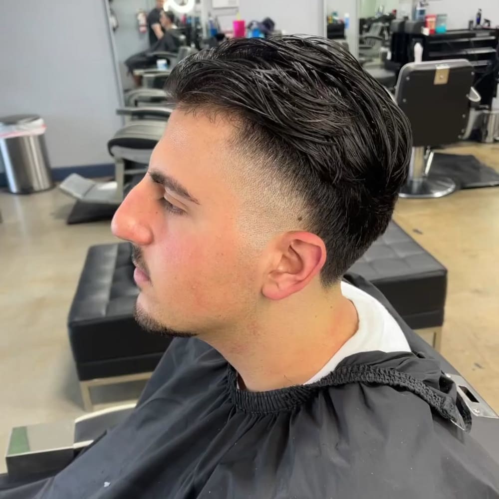 Man with straight hair styled with tapered sides and volume on top, featuring short, gradually faded sides and longer, slicked-back hair on top. He has a light mustache and goatee, wearing a black barber cape, sitting in a modern barbershop, showcasing a clean and contemporary look.