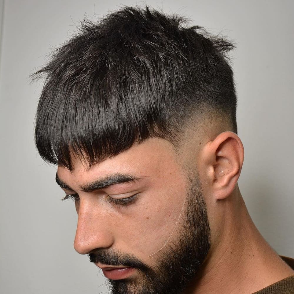 Man with straight hair styled in a thick fringe haircut, featuring a blunt fringe that falls over the forehead and short, faded sides. He has a well-groomed beard and is looking down, showcasing a modern and edgy look in a minimalist setting.