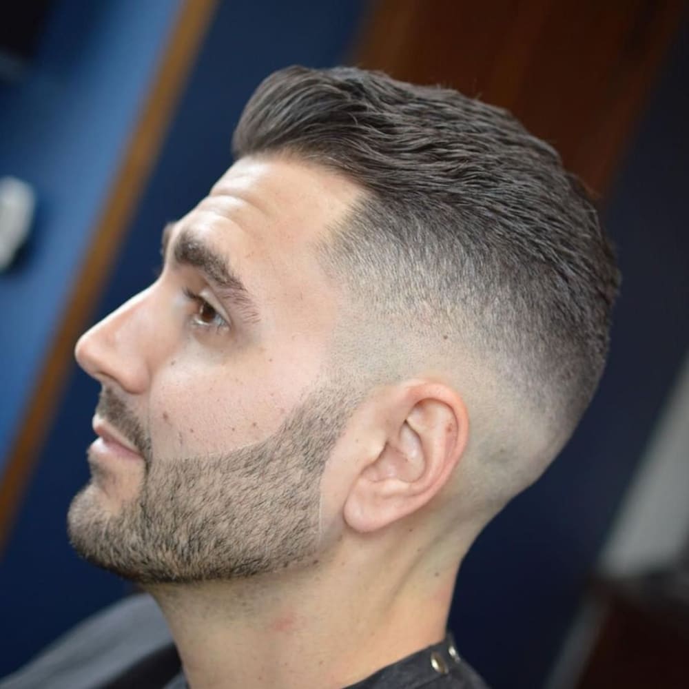 Man with straight, thick hair styled in a high fade haircut, featuring short sides that gradually blend into longer hair on top. He has a neatly groomed beard with sharp lines, sitting in a barbershop chair, showcasing a clean and modern look against a blue and wooden backdrop.