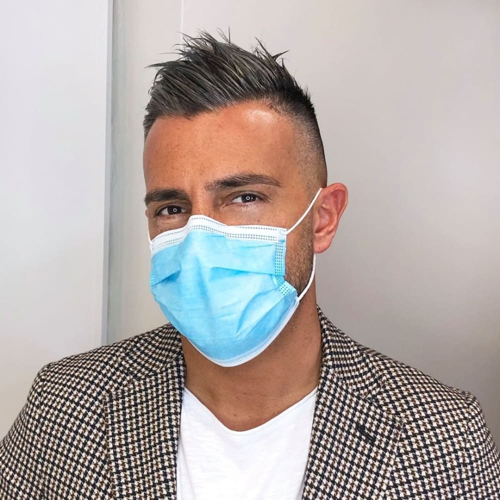 Man with straight hair styled in an undercut haircut, featuring short, closely shaved sides with longer, spiked hair on top. He is wearing a blue face mask and a checkered blazer over a white shirt, standing indoors against a neutral background, showcasing a stylish and professional look.