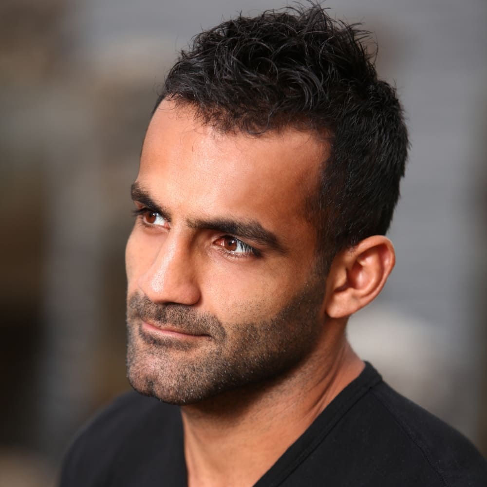 A close-up portrait of a man with a neatly groomed stubble beard. He has a strong, angular jawline, and his beard is trimmed to a short length, giving him a rugged yet polished appearance. His hair is styled short and slightly tousled, complementing the clean look of his beard. The man has a thoughtful expression, with deep-set eyes and a slight furrow in his brow. He is wearing a black shirt, which adds to the simplicity and sophistication of his overall look. The background is blurred, focusing attention on his facial features and beard style.