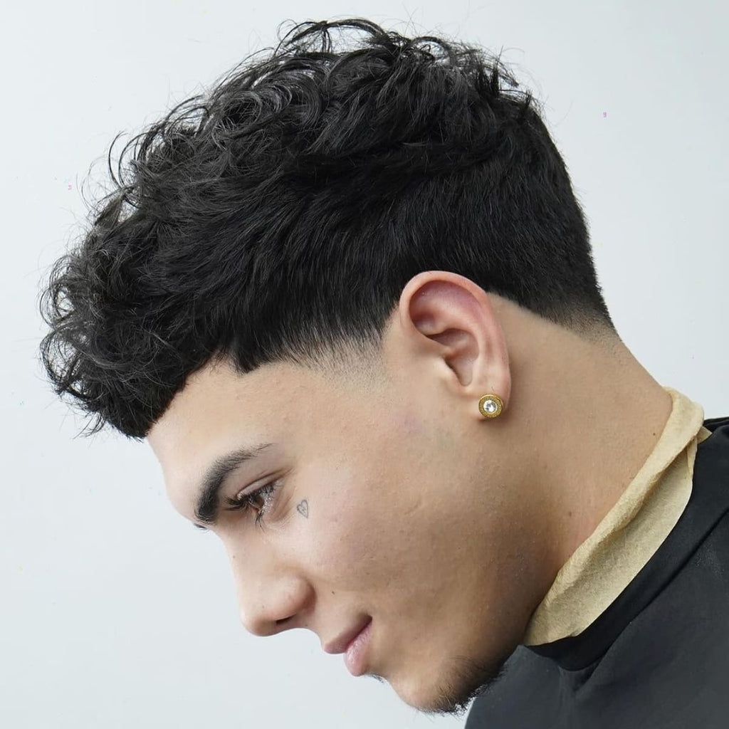 A side profile portrait of a young man with a fresh taper fade haircut and curly textured top. The hair on top is full and voluminous, with natural curls adding texture and dimension, while the sides are neatly tapered down, creating a smooth gradient. He wears a small, gold-studded earring and has a subtle tattoo under his eye, adding a stylish, edgy touch to his look. The background is a plain white, drawing full attention to the details of his haircut and facial features.