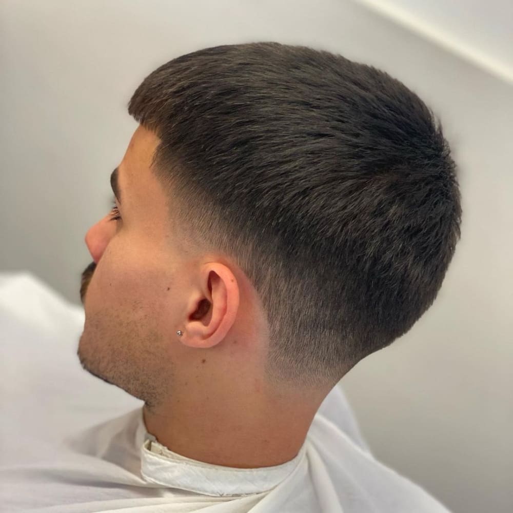 A close-up side profile of a man with a taper fade buzz cut hairstyle. The haircut features a longer length on top, gradually tapering down to a clean fade around the sides and back of the head. The top is left with a smooth, even texture, while the fade blends seamlessly into the skin, creating a sleek and polished look. The hairline at the forehead is softly rounded, and the overall style is neat and precise. The man is wearing a white barber's cape, and the background is blurred, keeping the focus on the sharp details of the taper fade. The haircut is modern and stylish, suitable for a clean, professional appearance.