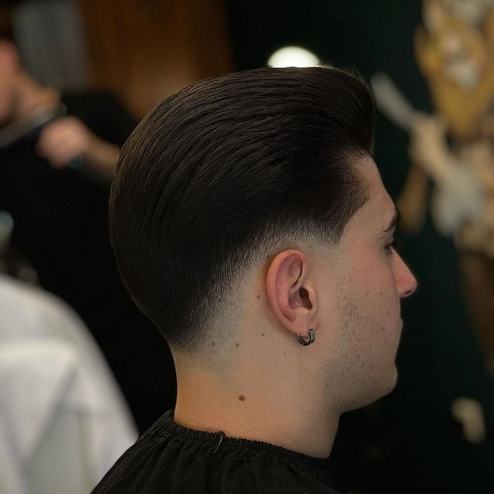 A man is seated in a barbershop, showcasing a sleek tapered pompadour haircut. The sides of his hair are gradually tapered down, creating a smooth transition from the longer top to the shorter sides. The top hair is styled back in a polished, voluminous manner, giving the classic pompadour look a modern touch. His hair appears thick and healthy, with a glossy finish that enhances the overall sophistication of the style. The barbershop background, slightly blurred, includes a barber at work, adding to the authentic atmosphere of the setting.