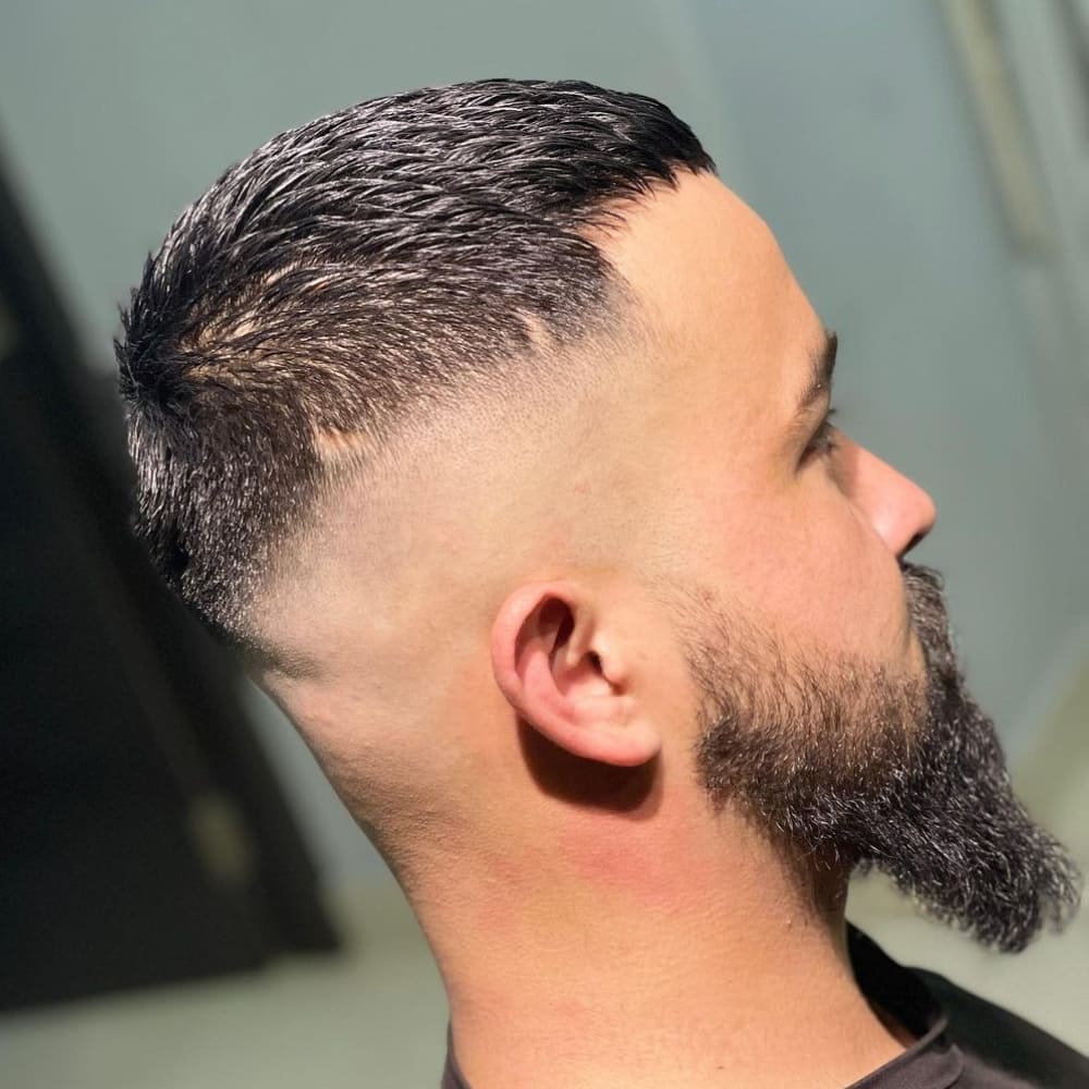 A close-up side profile of a man with a textured buzz cut fade hairstyle. The haircut features short, textured hair on top with visible layers that add dimension and movement. The sides and back are styled into a high skin fade, creating a sharp contrast with the thicker hair on top. The hairline is defined with a slight, straight edge at the forehead, and the overall look is both edgy and polished. The man also has a full, well-groomed beard that complements the clean lines of the fade, adding to the modern and stylish appearance. The setting is indoors, with the focus on the haircut’s detail and texture.