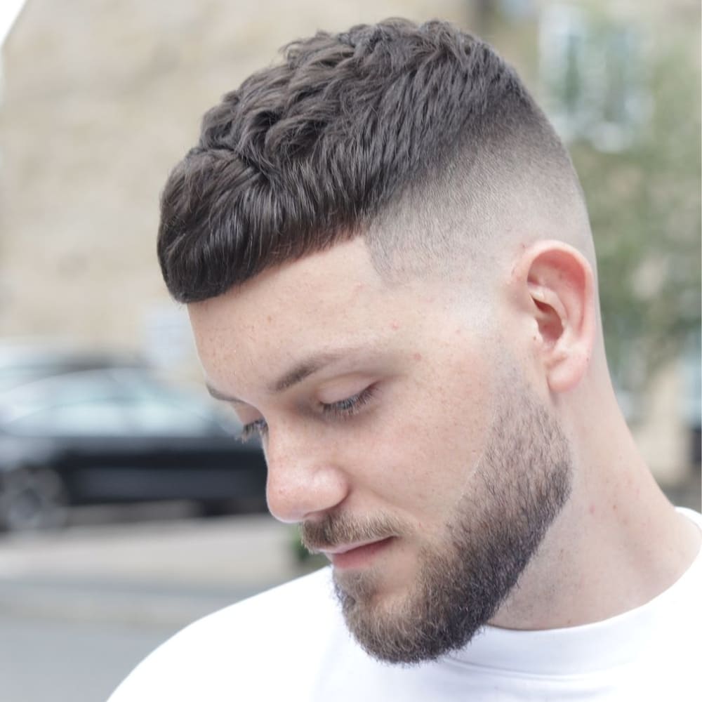 Man with a textured Caesar cut featuring a short, choppy fringe and a medium fade on the sides and back. The top hair is styled with natural waves for added texture and volume, complemented by a neatly trimmed beard, creating a fresh and contemporary look.