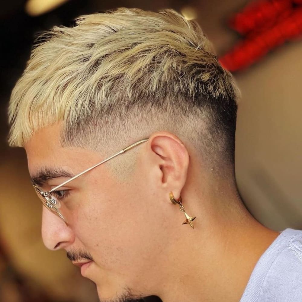 A man with a stylish textured crop fade haircut is depicted in the image. His hair features a textured, blonde top with choppy layers, creating a dynamic and voluminous look. The sides and back are faded down to the skin, offering a sharp and clean contrast. He is wearing gold-rimmed glasses and a distinctive star-shaped earring, which add to his fashionable appearance. The background suggests an indoor setting, possibly a barbershop or salon, highlighting the freshly done haircut.