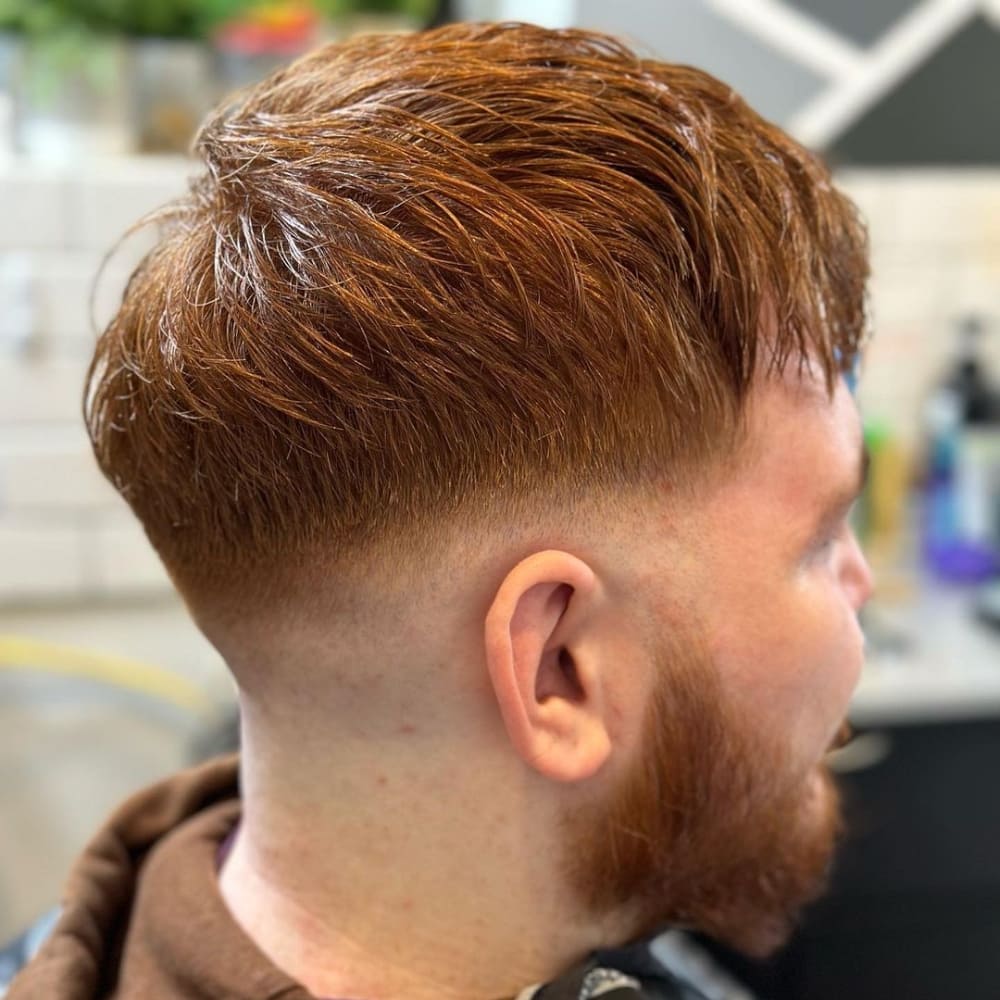 A man with a textured crop mid fade haircut is pictured in the image. His vibrant red hair is styled with a textured, layered top, adding depth and dimension to the look. The sides and back feature a mid fade, blending smoothly from longer hair on top to a shorter length around the ears and neckline. This creates a balanced and modern appearance. The man also sports a neatly trimmed red beard, complementing his hairstyle. The background suggests a barbershop setting, highlighting the fresh and professionally done haircut.