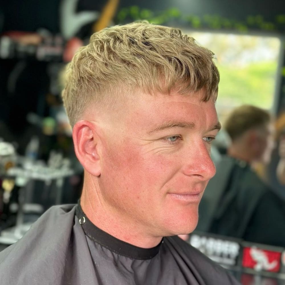 A man with a textured crop skin fade haircut is depicted in the image. His blonde hair is styled with a textured top, featuring choppy layers that add depth and movement. The sides and back are faded down to the skin, creating a sharp and striking contrast with the longer hair on top. This skin fade gives a clean, modern look. The man is seated in a barbershop, highlighting the fresh, professional cut. His expression is relaxed, showcasing the confidence and style that come with a well-executed textured crop hairstyle.