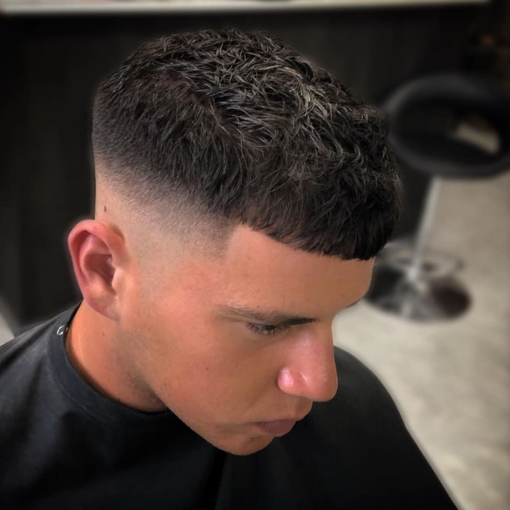 A young man with a fresh textured crop top fade haircut is shown in the image. The hairstyle features a short, textured top with neatly trimmed layers, creating a clean and stylish appearance. The sides and back are expertly faded, blending seamlessly from short to skin, adding a modern and sharp look. He is seated in what appears to be a barbershop, emphasizing the freshly done and precise nature of the haircut. The overall style highlights the natural texture of his hair while maintaining a well-groomed look.