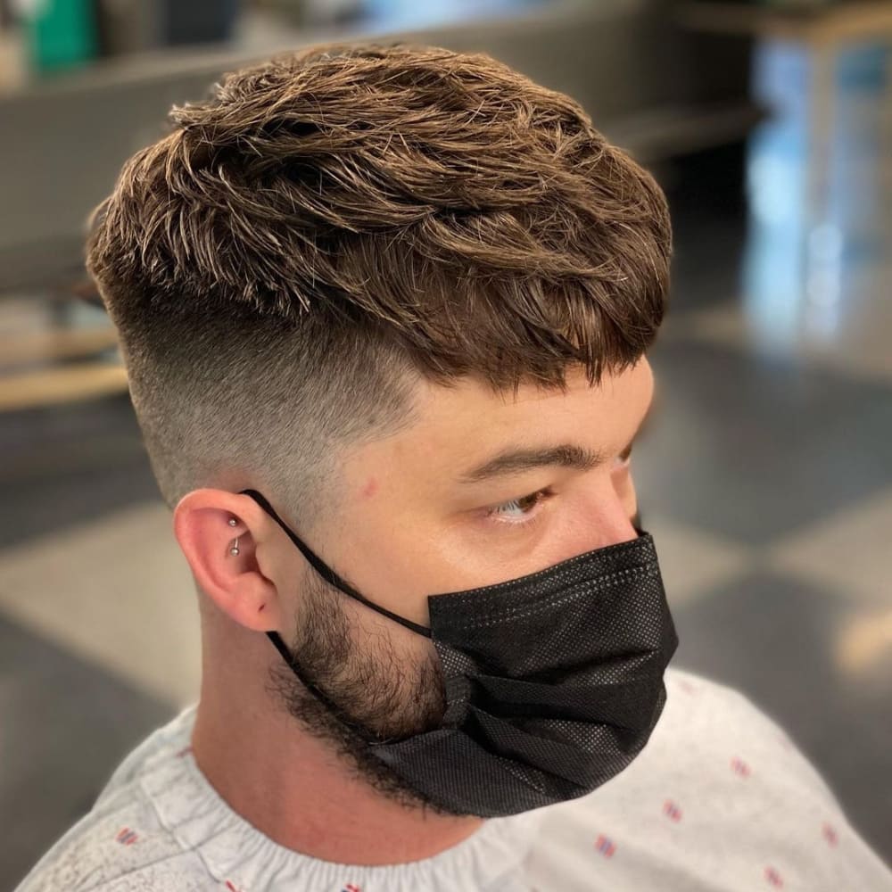 A man with a trendy textured French crop haircut is shown in the image. The hairstyle features a longer top with choppy, textured layers, adding volume and movement, while the sides and back are faded short, creating a clean and modern contrast. He is wearing a black face mask and has a small ear piercing. The setting appears to be a barbershop, emphasizing the freshly styled look.