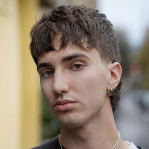 A young man is shown with a textured fringe haircut, characterized by choppy, layered bangs that fall naturally across his forehead, creating a casual, tousled look. His hair is dark brown, with a slightly longer length at the top that adds volume and movement, while the sides are tapered and blend seamlessly into a subtle mullet at the back. He has a soft, contemplative expression, with piercing green eyes and well-groomed eyebrows. The man is wearing a simple silver hoop earring in his left ear, which complements his minimalistic style. He is photographed outdoors, with a blurred background featuring muted colors, possibly from urban surroundings, which further emphasizes his relaxed and modern appearance.