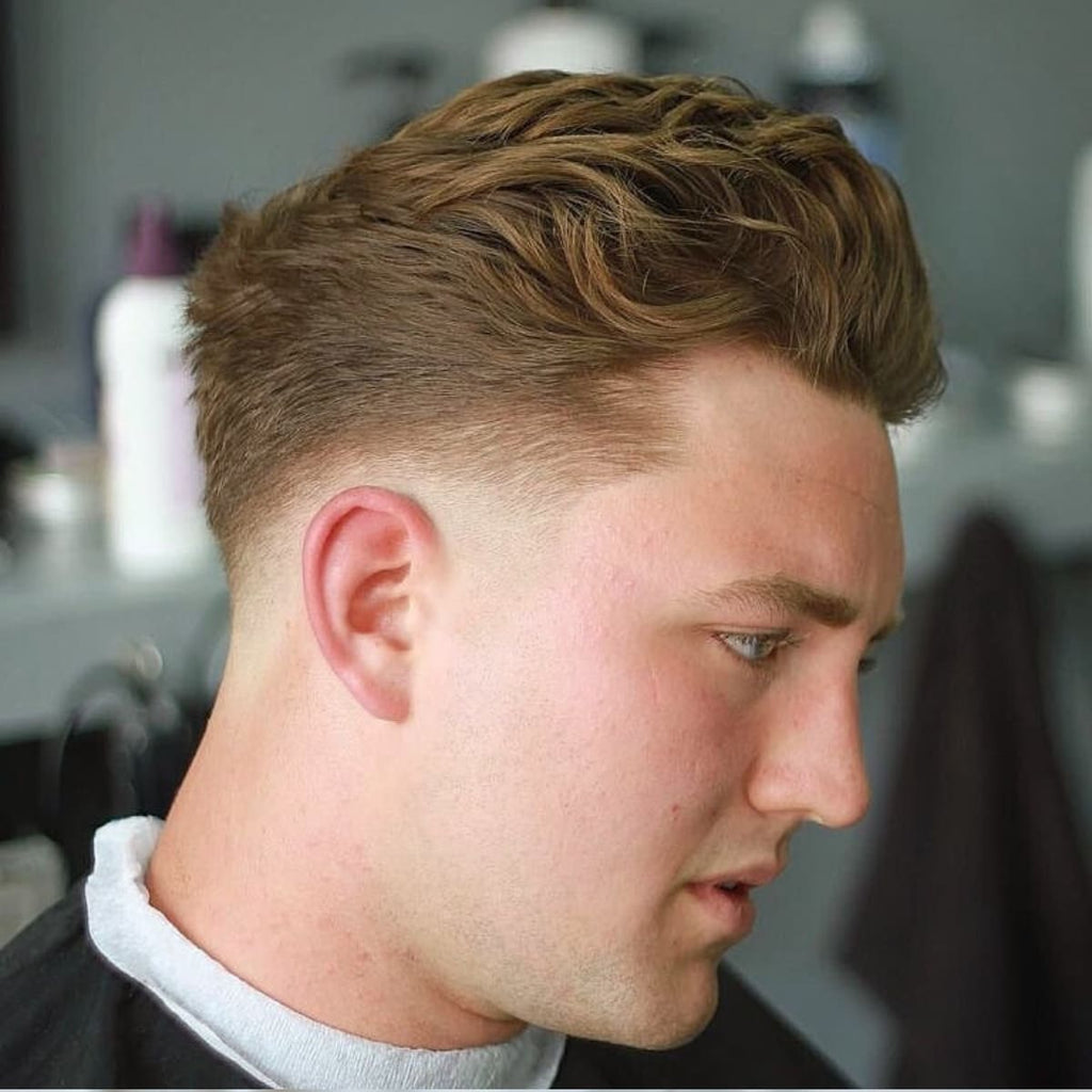 A side profile of a man with a textured low fade slick back haircut, showcasing a voluminous, layered top styled with natural texture and a relaxed finish. The low fade starts at the base of the neckline and tapers gradually below the ears, creating a subtle and clean transition that emphasizes the fullness of the textured top. The haircut has a modern and casual vibe, perfect for versatile styling. The background features a barbershop setting with muted tones and styling products visible on a shelf, keeping the focus on the detailed haircut.