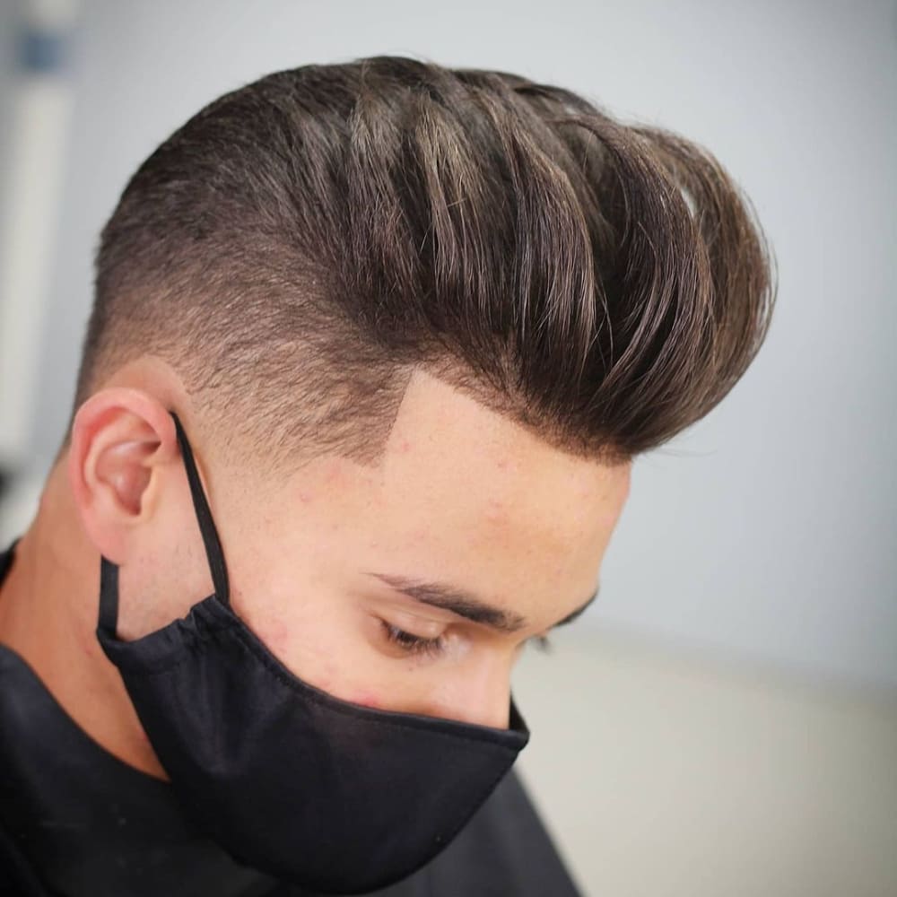 A man is shown with a stylish textured pompadour haircut. His hair is voluminous and styled upwards with a natural, slightly messy texture, adding depth and dimension. The sides are neatly faded, creating a clean, sharp contrast with the top. He is wearing a black face mask, partially covering his face but allowing the focus to remain on the meticulously styled hair. The setting is a modern barbershop, highlighting the precision and care taken to achieve this contemporary look. The overall effect is a fashionable, effortlessly chic pompadour that exudes confidence and style.