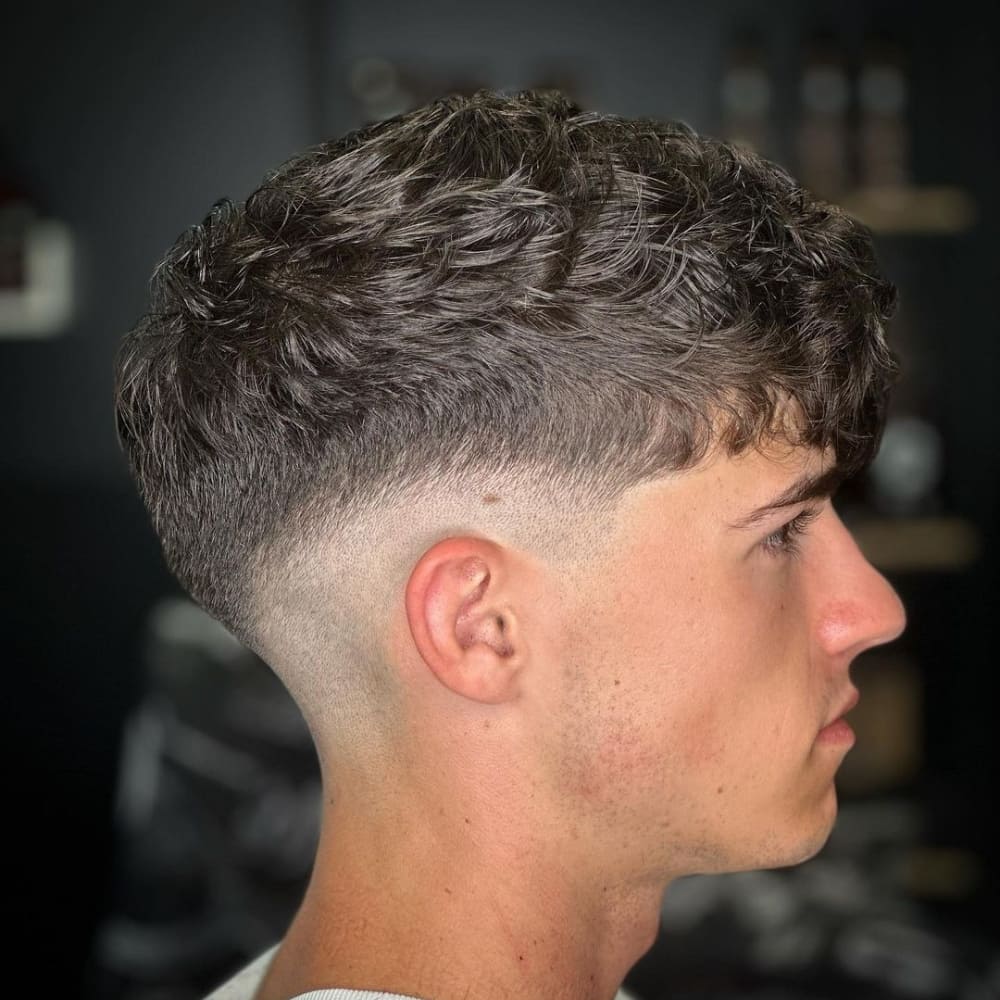 A young man with a textured hairstyle featuring a mid fade. The hair on top is styled with natural waves, providing a casual and trendy appearance. The mid fade begins around the temple area, blending smoothly down to the skin at the base of the neck and around the ears. The clean lines of the fade accentuate the texture and volume of the hair on top. The man is viewed from the side, showcasing the sharp contrast between the longer hair on top and the closely cropped fade. The background is slightly blurred, drawing focus to the stylish haircut.