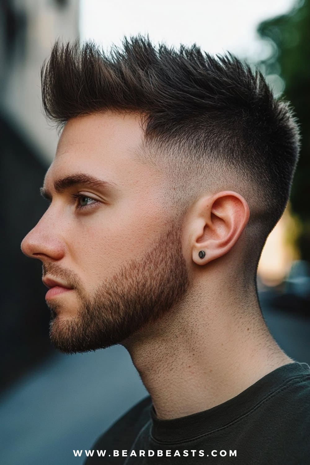 A young man with striking facial features is sporting a Faux Hawk, a bold and edgy choice for men. His hair is styled with a pronounced lift in the center, creating the signature faux hawk look, while the sides are sharply faded, enhancing the contrast and giving the hairstyle a modern, clean-cut appeal. The textured top adds volume and height, making this style ideal for those who want to stand out with a daring yet wearable haircut. His beard is well-groomed, complementing the angular lines of the faux hawk and adding to his overall sharp, confident appearance.