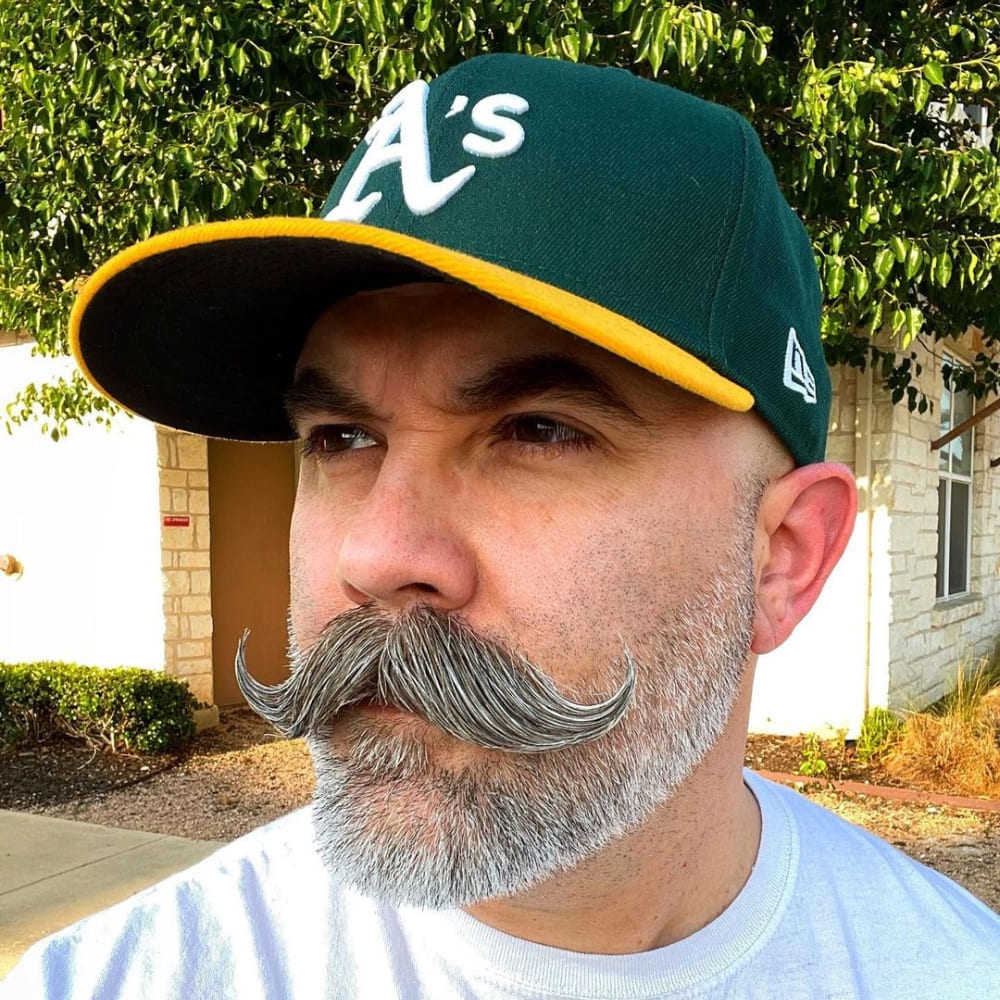 A man displaying a handlebar beardstache, featuring a prominent handlebar mustache with curled ends and a neatly trimmed short beard. He is wearing a green and yellow A's baseball cap and a white t-shirt. The background includes greenery and a building, enhancing the vintage and distinguished appeal of the handlebar beardstache style. The overall look is both classic and stylish, perfectly capturing the essence of this facial hair trend.