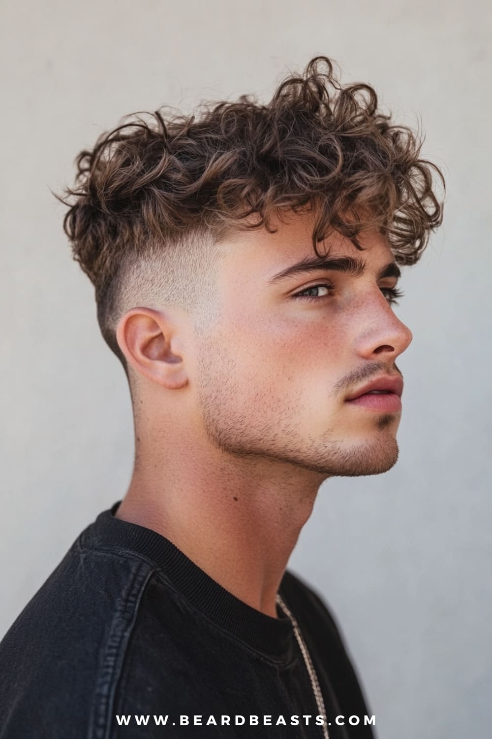 A young man with a defined jawline is showcasing a Medium-Length Undercut, a trendy and popular choice among medium length hairstyles for men. His hair features a voluminous top with natural curls that add texture and movement, while the sides are sharply faded, creating a striking contrast that emphasizes the height and fullness of the top. The curls are styled in a loose, effortless way, giving the hairstyle a casual yet polished look. This undercut is perfect for those who want to maintain a stylish and modern appearance without sacrificing the natural texture of their hair. He is dressed in a black shirt, with a silver chain peeking out, complementing his contemporary style.