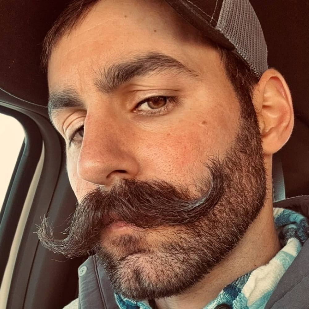 A man with a scruffy beardstache, characterized by a thick, curled mustache and a slightly longer, rugged beard. He is wearing a baseball cap and a checkered shirt, seated inside a car. The casual and adventurous look is enhanced by the natural, unrefined texture of his facial hair, perfectly embodying the carefree and effortless style of the scruffy beardstache. The overall appearance is rugged and stylish, capturing the essence of this popular facial hair trend.