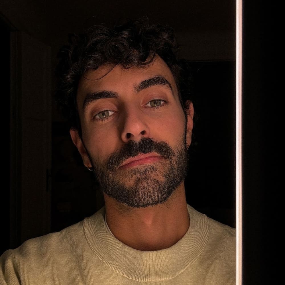 A man with a subtle beardstache, featuring a neatly trimmed mustache and a closely cropped beard. He has curly dark hair and is wearing a light beige sweater. The lighting highlights his facial features and the understated elegance of his beardstache. The overall look is refined and sophisticated, perfectly capturing the minimalistic and professional style of the subtle beardstache.