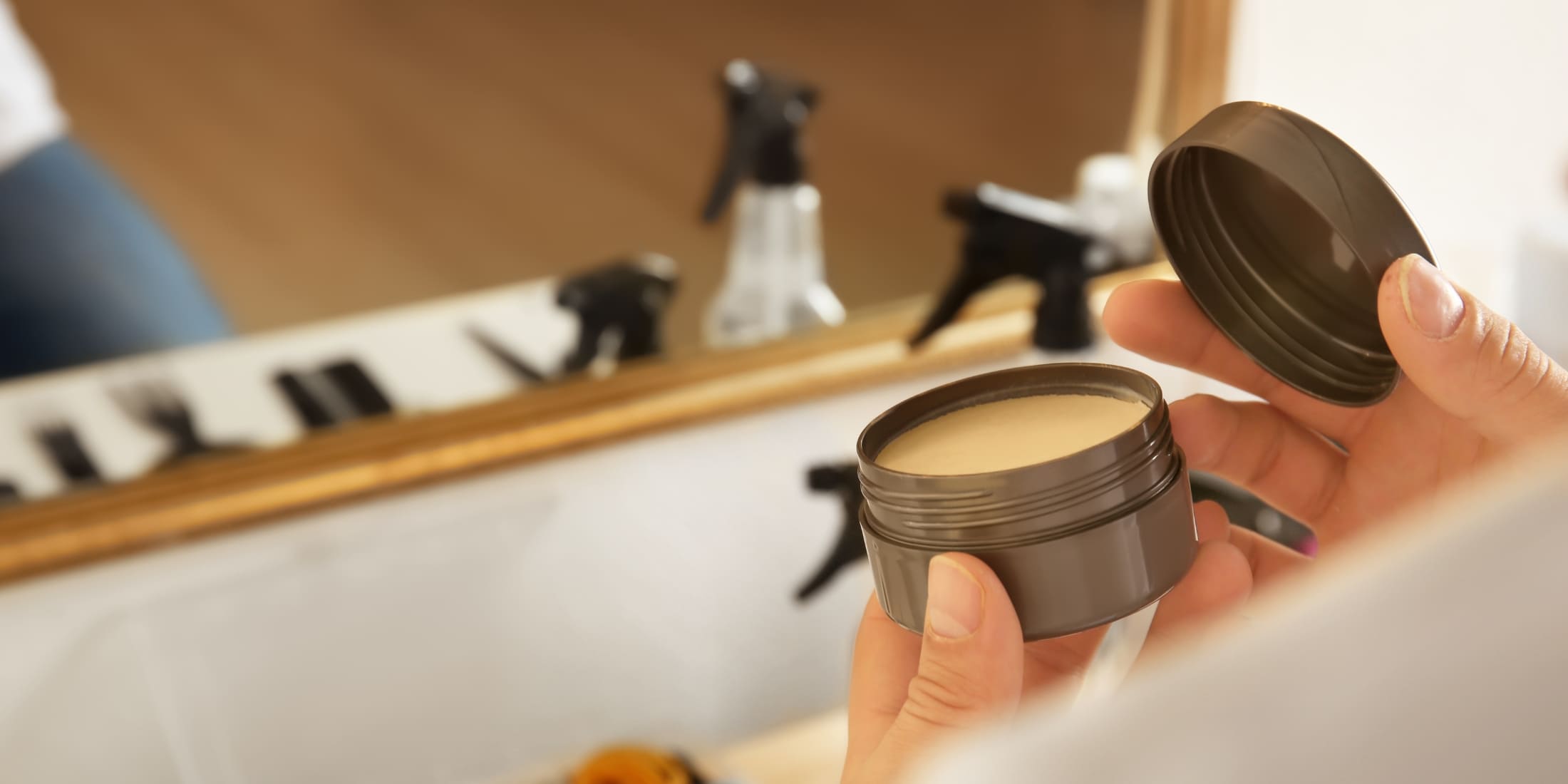 Man opening a black tin of pomade in a well-lit barber shop, reflecting in a mirror with hair styling tools in the background, illustrating a guide on how to use pomade on curly hair.