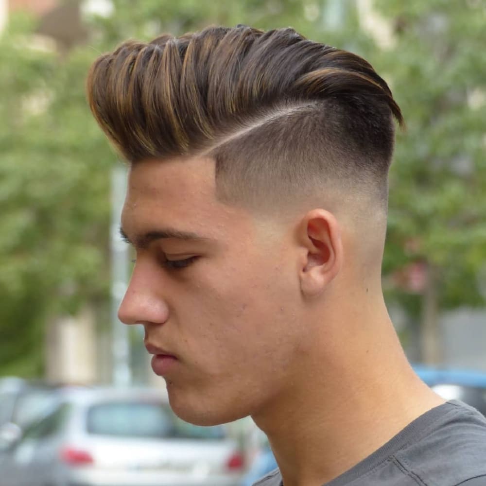 A young man with an undercut pompadour haircut is standing outdoors. His hairstyle features a sharply defined undercut with the sides closely shaved, while the top hair is voluminous and swept back in a classic pompadour style. The hair has subtle highlights, adding depth and texture to the look. The sharp parting line adds a touch of precision and modernity. His expression is serious, and he is wearing a casual grey t-shirt, contrasting with the stylish and well-groomed haircut. The background is slightly blurred, showing a street scene with greenery and parked cars, emphasizing the focus on his striking hairstyle.