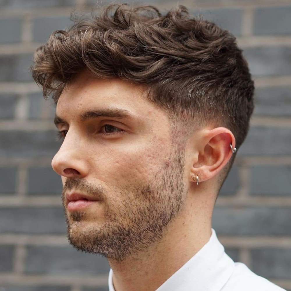 Man with a wavy Caesar haircut featuring textured, wavy hair on top and a tapered fade on the sides and back. He has a light stubble beard and is wearing small hoop earrings in both ears. Dressed in a white shirt, he stands against a brick wall, highlighting the natural waves and volume of his hair.