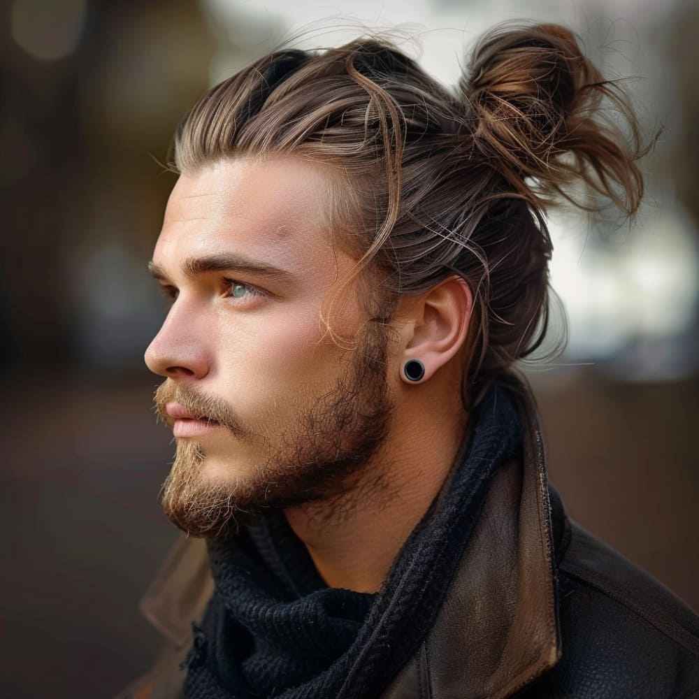 A man with a stylish wavy man bun is featured in this image, showcasing his effortlessly cool and modern look. His long, wavy hair is pulled back into a messy yet intentional bun at the back of his head, with a few loose strands adding texture and a relaxed vibe. The sides are neatly tucked behind his ears, revealing a subtle undercut and a pair of black ear gauges, which add an edgy touch to his appearance. His well-groomed beard complements the hairstyle, enhancing his rugged, masculine look. Dressed in a dark scarf and a leather jacket, he exudes a sense of confidence and individuality. The soft, diffused lighting highlights the natural waves in his hair and the rich tones of his complexion, creating a warm and inviting atmosphere. The image captures a perfect blend of casual style and contemporary edge.