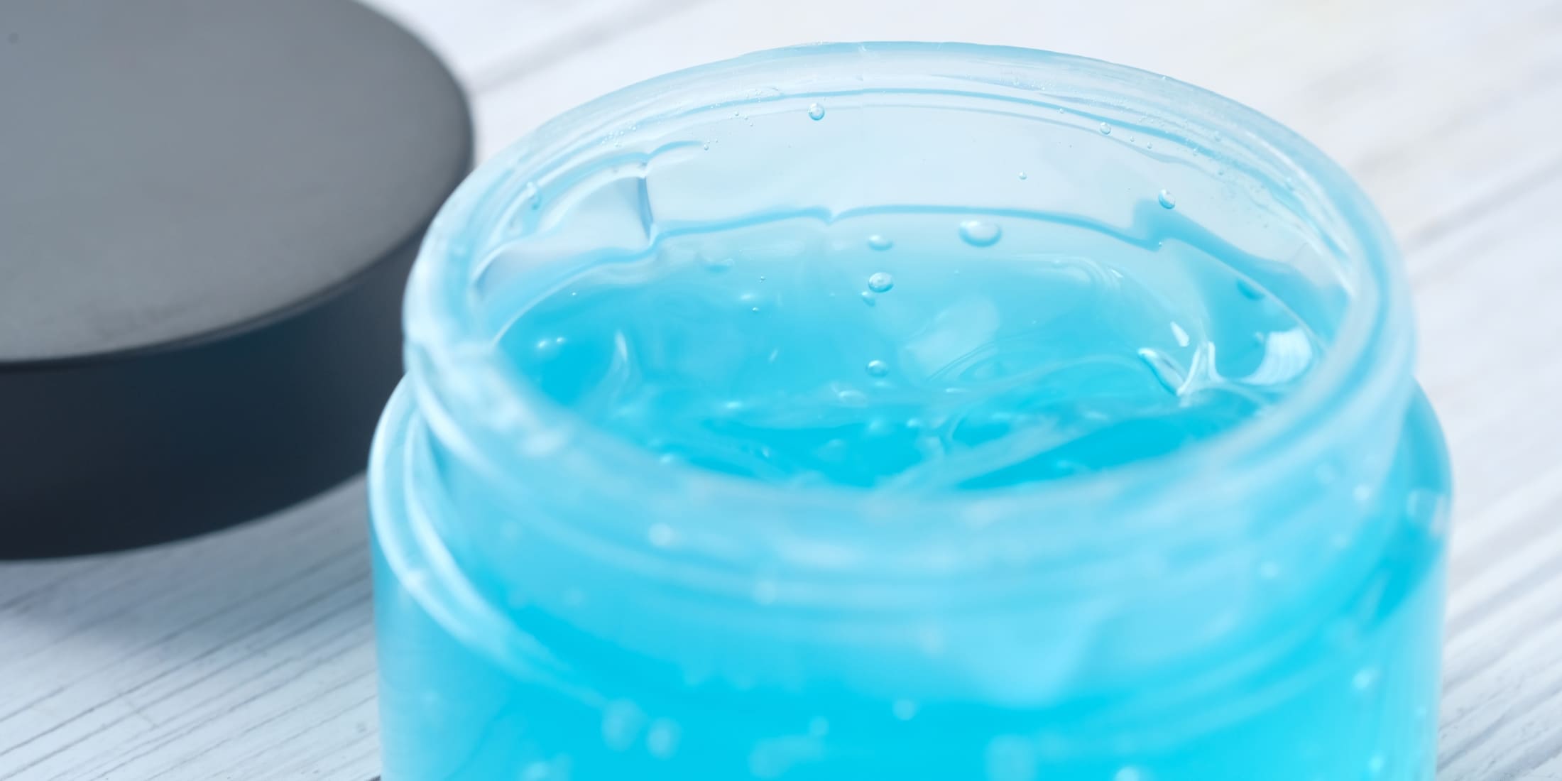 An open jar of blue hair gel, showcasing its smooth, shiny texture. The jar lid is placed beside it on a light-colored wooden surface. This image visually represents what is hair gel—a versatile hairstyling product used to achieve various looks.