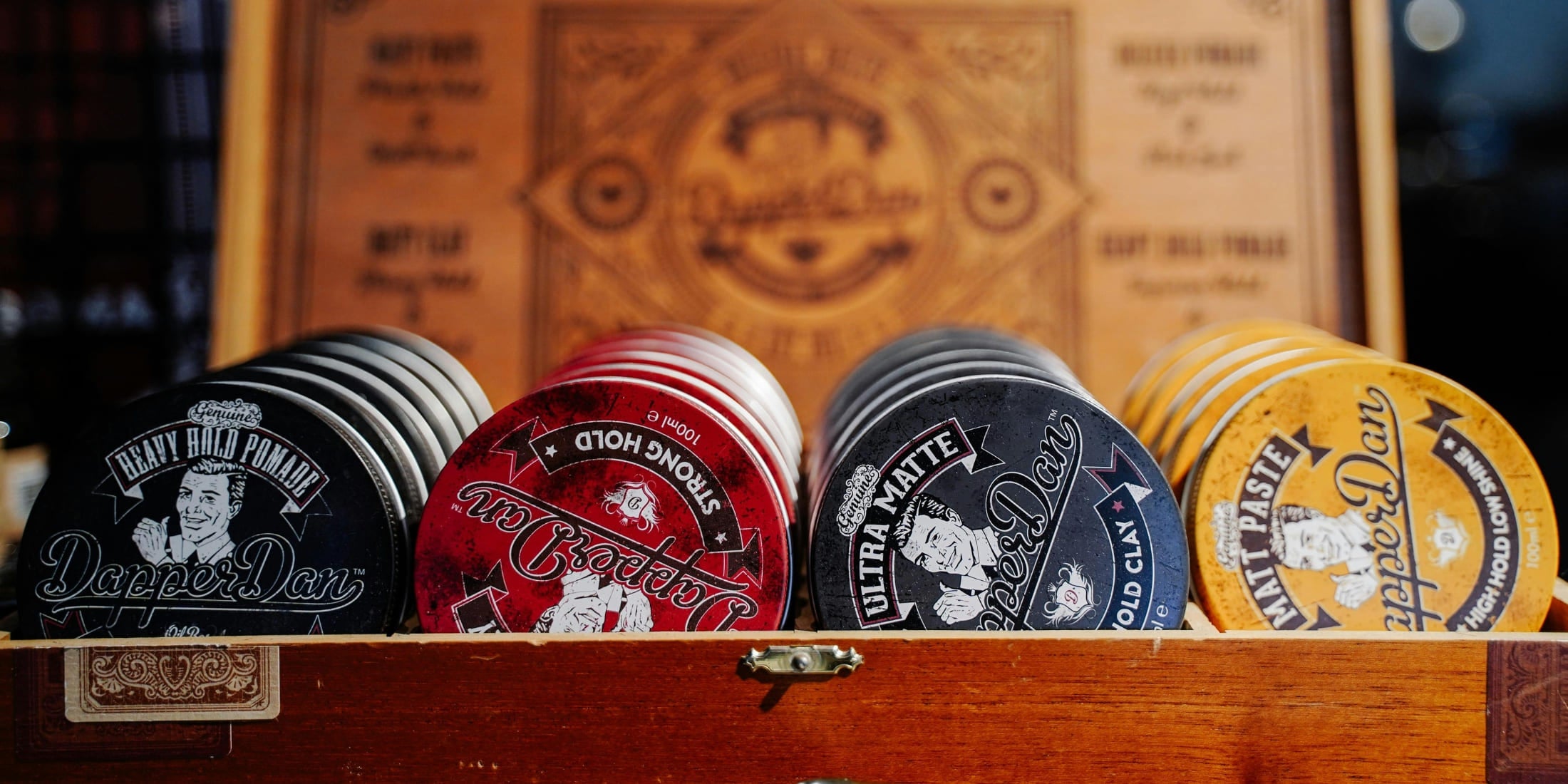 A close-up image of various hair styling products displayed in a wooden box. The products include tins of heavy hold pomade, strong hold, ultra matte, and matt paste from the brand Dapper Dan. The background features a vintage-style sign with branding details, creating a nostalgic barbershop ambiance. The products are neatly arranged, showcasing different types of hair paste and pomade options available for various styling needs.